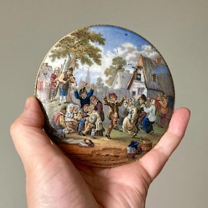 Puede incluir: Un plato decorativo antiguo, pequeño y redondo, con una ilustración detallada y colorida de una escena de pueblo. El plato presenta un borde dorado y una imagen pintada de gente bailando y tocando música. El texto "THE VILLAGE WEDDING" es visible.