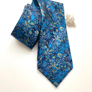 Pode incluir: Uma gravata com fundo azul escuro e um padrão repetitivo de folhas, vinhas e formas florais em azul claro, verde-azulado e verde-oliva. A gravata está dobrada sobre si mesma e posicionada ao lado de uma pequena concha branca em espiral.