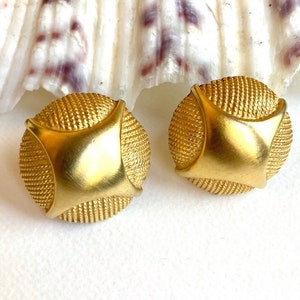 Puede incluir: Un par de pendientes de color dorado con un diseño circular texturizado. Los pendientes tienen un centro cuadrado elevado.