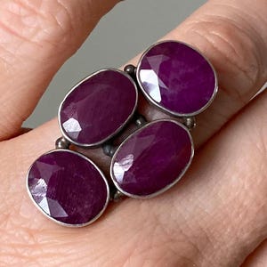 Könnte beinhalten: Ein Silberring mit vier ovalen, tiefmagentafarbenen Edelsteinen. Die Edelsteine sind in einzelnen Fassungen gefasst und in einem Cluster-Design angeordnet. Der Ring wird an einem Finger getragen.