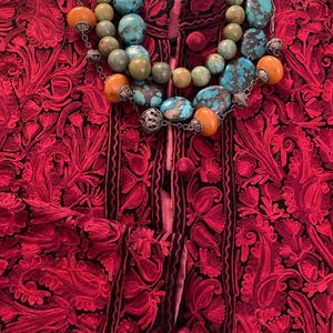 May include: A red embroidered jacket with black trim and a turquoise, amber, and silver beaded necklace. The jacket features a detailed floral pattern and small, round buttons. The necklace has large, round beads.