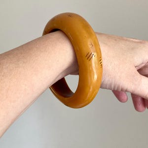 Pode incluir: Uma pulseira tipo bracelete grossa e arredondada em uma cor amarelo-mostarda sólida. A superfície do bracelete apresenta pequenas marcas lineares escuras gravadas distribuídas pelo exterior, criando uma aparência texturizada no material liso e polido.