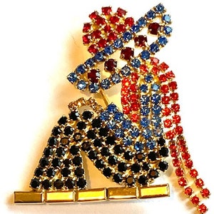 May include: A rhinestone brooch featuring a seated figure with a red, blue, and black design. The figure is wearing a hat and has a long, flowing train.
