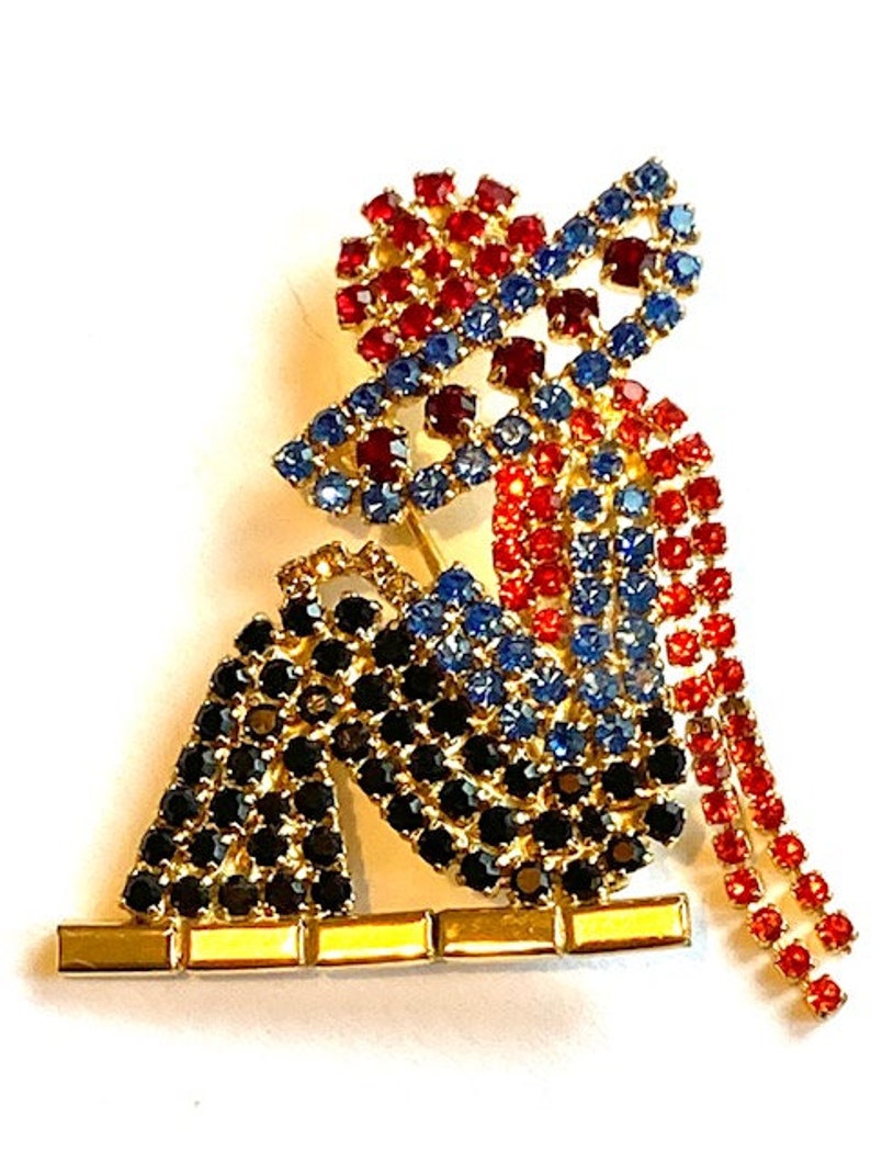 May include: A rhinestone brooch featuring a seated figure with a red, blue, and black design. The figure is wearing a hat and has a long, flowing train.
