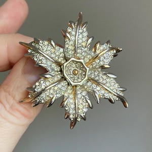 May include: A vintage brooch in the shape of a flower with multiple petals. The brooch is silver-toned with clear, sparkling rhinestones covering the petals. The center of the flower has a gold-toned, circular design.