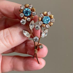 May include: A vintage floral brooch featuring two flower blooms with light blue center stones and clear crystal accents. The gold-toned metal brooch has a stem and leaf design, with a cluster of crystals at the base.