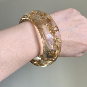 Puede incluir: Un brazalete grueso y transparente con inclusiones de pan de oro. El brazalete de resina transparente tiene forma redondeada y se lleva en la muñeca. El pan de oro añade un toque de lujo al accesorio.