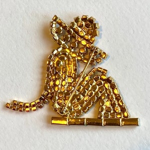 May include: A gold-toned brooch depicting a seated figure with a hat, made of small, sparkling rhinestones. The figure is holding a stick in its hand, and the brooch has a pin back.