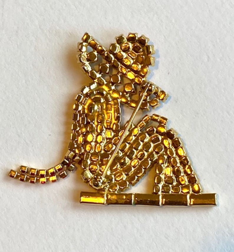 May include: A gold-toned brooch depicting a seated figure with a hat, made of small, sparkling rhinestones. The figure is holding a stick in its hand, and the brooch has a pin back.