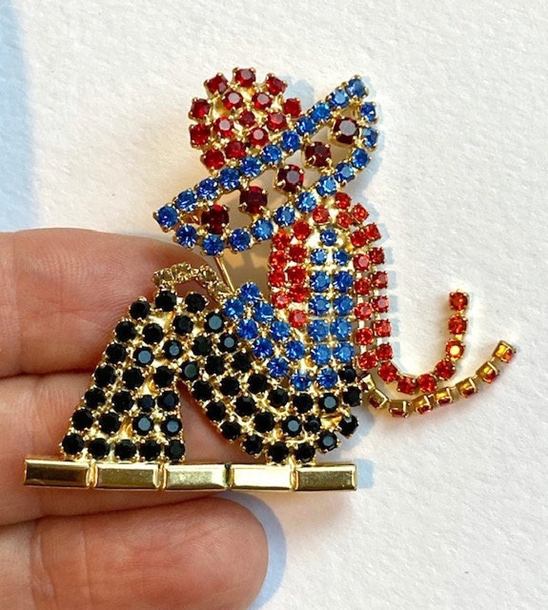 May include: A gold-toned brooch in the shape of a monkey, adorned with red, blue, and black rhinestones. The monkey is sitting with its legs crossed and its tail curled around its body.