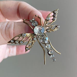 May include: A vintage dragonfly brooch with a gold-tone metal frame and clear crystal accents. The dragonfly has detailed wings and a large central crystal. The brooch is held up by a hand.