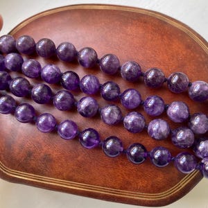 May include: Four strands of amethyst gemstone beads. The beads are a deep purple color and appear to be polished. The strands are resting on a brown, wooden surface. The beads are round and appear to be strung together.