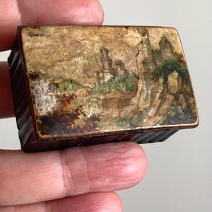 May include: A small rectangular box with a dark brown, ribbed base and a lid featuring a weathered, painted landscape scene. The artwork depicts a castle or stone structure set against a muted, textured background with earthy tones of tan, green, and brown.