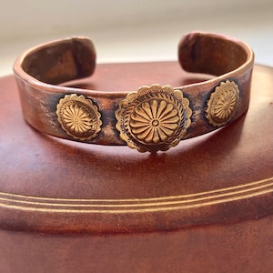 Puede incluir: Una pulsera de puño de cobre con tres elementos decorativos circulares de color dorado. La pulsera tiene un aspecto rústico y envejecido. Los elementos dorados presentan un diseño de rayos de sol. La pulsera se muestra sobre una superficie marrón.