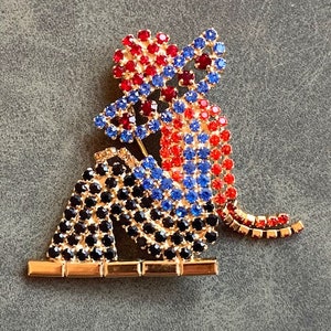 May include: A red, blue, and black rhinestone brooch depicting two figures sitting on a gold bar. The figures are wearing hats and have a whimsical, cartoon-like style.