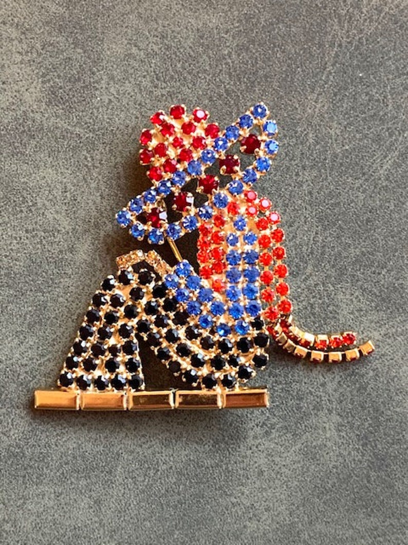 May include: A red, blue, and black rhinestone brooch depicting two figures sitting on a gold bar. The figures are wearing hats and have a whimsical, cartoon-like style.