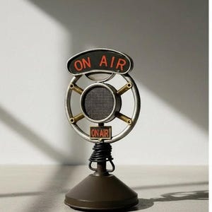 Puede incluir: Un letrero decorativo de metal con forma de micrófono que presenta un marco circular con un elemento de altavoz de malla central. El letrero muestra el texto ON AIR en letras naranjas sobre un fondo negro en la parte superior y un letrero ON AIR más pequeño.