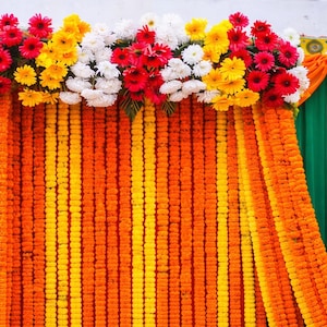 K&ouml;nnte beinhalten: Ein dekorativer Hintergrund mit vertikalen Str&auml;ngen aus orangefarbenen und gelben Ringelblumen. Die obere Kante ist mit einem dichten Arrangement aus roten, gelben und wei&szlig;en Gerbera-Bl&uuml;ten verziert, flankiert von drapierten orangefarbenen und gr&uuml;nen Stoffbahnen.