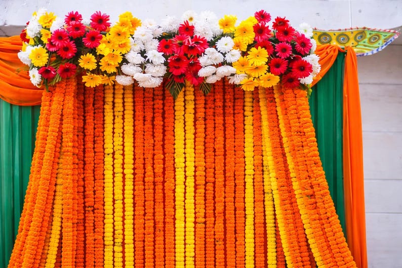 K&ouml;nnte beinhalten: Ein dekorativer Hintergrund mit vertikalen Str&auml;ngen aus orangefarbenen und gelben Ringelblumen. Die obere Kante ist mit einem dichten Arrangement aus roten, gelben und wei&szlig;en Gerbera-Bl&uuml;ten verziert, flankiert von drapierten orangefarbenen und gr&uuml;nen Stoffbahnen.