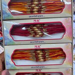 May include: Four rectangular boxes containing decorative wrist bands, each featuring red and yellow threads, gold-toned accents, and central light-coloured oval beads. The boxes are labelled with the text M.R Mahesh Rakhi and Ek Anokhi Pehchan.