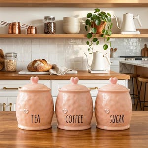 May include: Three pink ceramic kitchen canisters with heart-shaped lids and small heart details. The canisters are labelled "TEA", "COFFEE", and "SUGAR" in black script. The textured surface adds a rustic charm to the kitchen decor.