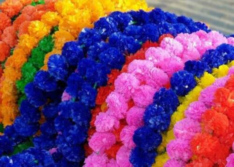 K&ouml;nnte beinhalten: Eine Ansammlung k&uuml;nstlicher Blumengirlanden mit dichten, ger&uuml;schten Bl&uuml;tenbl&auml;ttern in leuchtenden Farben wie Blau, Pink, Gelb, Orange, Rot und Gr&uuml;n. Die Str&auml;nge sind in parallelen Reihen angeordnet und bilden ein buntes Arrangement aus Kunstblumen.