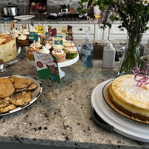 Op de afbeelding: Een tafel met een verscheidenheid aan desserts, waaronder een cheesecake met een roze "It's a Girl"-topper, cupcakes met kleurrijke sprinkles, koekjes en een trifle. Er staat ook een boek met de titel "Als je een muis een koekje geeft" op een standaard.