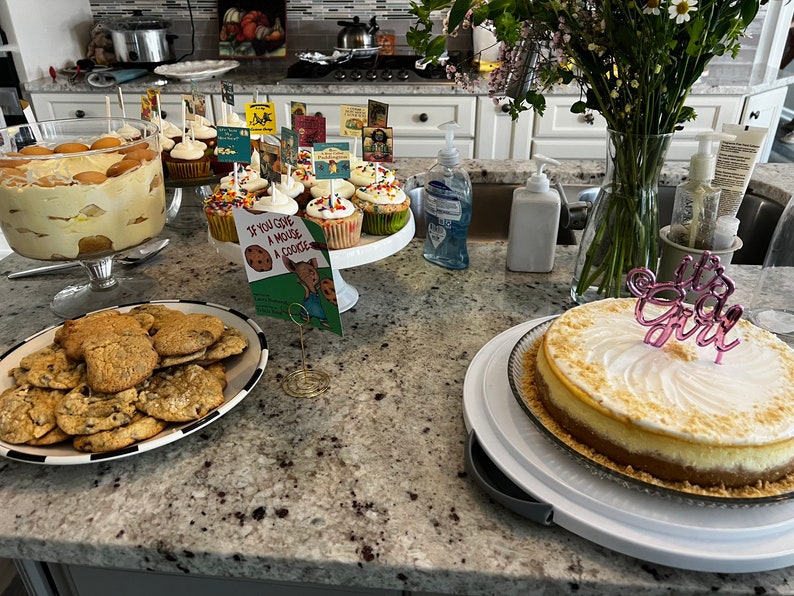 Op de afbeelding: Een tafel met een verscheidenheid aan desserts, waaronder een cheesecake met een roze "It's a Girl"-topper, cupcakes met kleurrijke sprinkles, koekjes en een trifle. Er staat ook een boek met de titel "Als je een muis een koekje geeft" op een standaard.