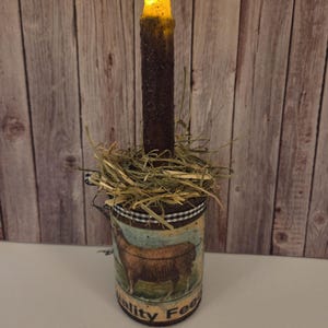 May include: A rustic candle holder with a flickering LED candle. The candle is dark brown with a yellow flame. The base is a tin can decorated with a goat illustration and the words "Quality Feed". The candle is surrounded by dried grass.