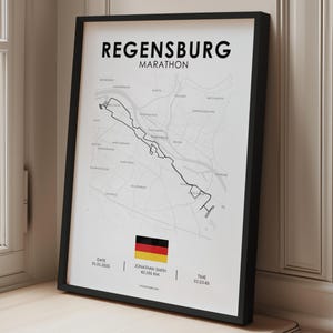 Puede incluir: Un póster enmarcado que muestra un mapa de la ruta del Maratón de Ratisbona en líneas negras sobre fondo blanco. El texto dice Regensburg Marathon, Jonathan Smith, 42,195 km, fecha 01.01.2025 y tiempo 01:23:45, con un icono de la bandera alemana.