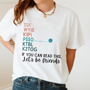 May include: White t-shirt with the text "SSK WYIB K1P1 PSSO KTBL K2TOG IF YOU CAN READ THIS, Let's be friends." The shirt features a teal yarn ball graphic. The shirt is worn with blue jeans.
