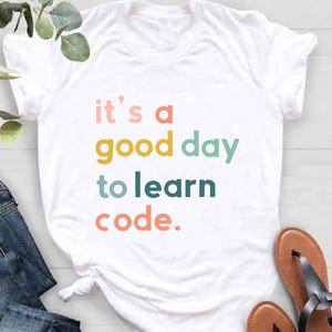 May include: White t-shirt with the text "it's a good day to learn code" in colorful letters. The shirt is paired with blue jeans and brown sandals. The image is taken from a top-down perspective.