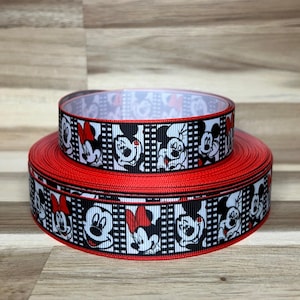 May include: Two stacked rolls of grosgrain ribbon featuring a repeating pattern of Mickey and Minnie Mouse faces in black, white, and red. The design includes vertical black and white film strip borders between the character illustrations.