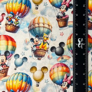 May include: Fabric featuring a repeating pattern of Disney characters in hot air balloons. Mickey Mouse, Minnie Mouse, Donald Duck, and Pluto are seen in colorful balloons against a cloudy sky background. The balloons have rainbow and striped designs.