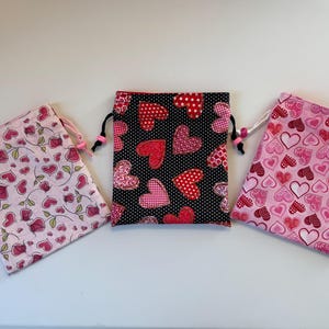 May include: Three small fabric drawstring bags with pink and red heart patterns. The bags are made of cotton and have a drawstring closure. The bags are perfect for small gifts or favors.