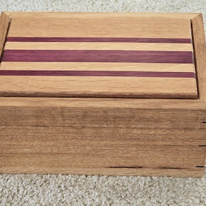 May include: A wooden box with a hinged lid. The lid is made of light wood with three thin strips of darker wood inlaid into the surface. The box is made of the same light wood.