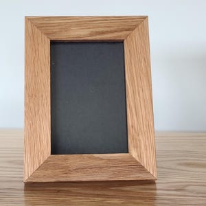 May include: A rectangular picture frame featuring a light brown wood grain finish with mitered corners. The frame surrounds a solid black rectangular center panel, creating a clean and simple display piece for photographs or artwork.