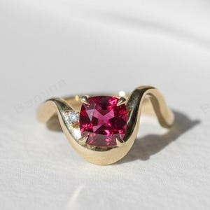 Puede incluir: Un anillo de oro con una gema roja oscura de talla cojín como pieza central. El anillo tiene un diseño único en forma de ola con una pequeña piedra de acento transparente en un lado. Sobre un fondo blanco.