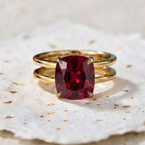 May include: A gold ring featuring a large, cushion-cut deep red gemstone set in the centre. The band consists of two parallel gold wires that meet at the setting, creating a layered appearance around the vibrant, faceted red stone.