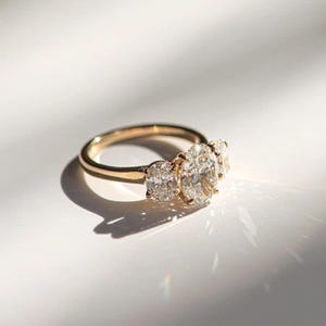 May include: A gold ring featuring three oval-cut diamonds. The central diamond is larger than the flanking stones. The ring sits on a white surface, with shadows creating depth. The ring's band is a simple, polished gold.