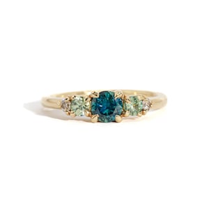 Può includere: Un anello con fascia in oro con una pietra centrale di zaffiro blu e pietre preziose verdi e bianche più piccole.