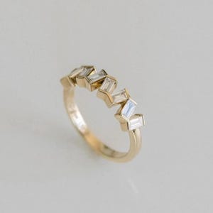 Puede incluir: Un anillo de oro con un diseño en zigzag de piedras preciosas rectangulares y transparentes. El anillo tiene una superficie pulida y está sobre un fondo suave y neutro. Las piedras están espaciadas uniformemente.