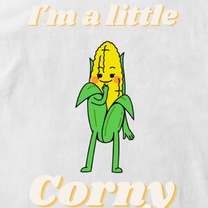 Op de afbeelding: Een wit T-shirt met een gele en groene cartoonmais met de tekst "I'm a little corny" in geel.