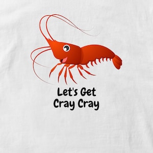 Puede incluir: Camiseta blanca con un camarón de dibujos animados en tonos rojos y naranjas. El camarón tiene una cara sonriente y largas antenas. Debajo del camarón está el texto "Let's Get Cray Cray" en negro.