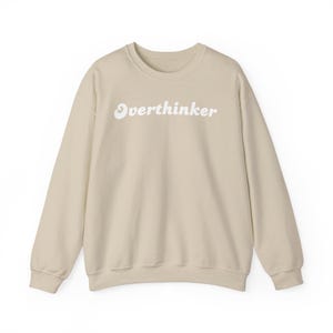 Op de afbeelding: Een beige sweatshirt met het woord "Overthinker" in wit op de voorkant.