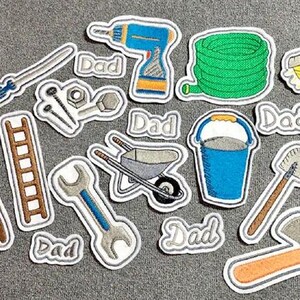 May include: A set of iron-on patches featuring tools and the word "Dad". The patches include a drill, a wrench, a ladder, a shovel, a wheelbarrow, a bucket, a hose, a broom, an axe, and a set of nuts and bolts.