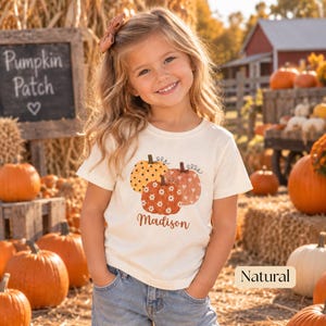 May include: A cream-colored short-sleeve t-shirt featuring a graphic of three pumpkins with polka dot, daisy, and heart patterns, with the name Madison printed below in brown script. The shirt is paired with blue denim jeans.