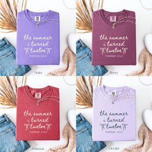 May include: Four t-shirts in violet, berry, crimson, and orchid colors, each folded and displaying the text "the summer i turned twelve VINTAGE 2014". Each shirt has a necklace and is paired with jeans and a neutral-colored scarf.