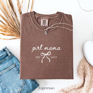 May include: An espresso-colored short-sleeve t-shirt featuring the white text girl mama, a graphic of a bow, and the text EST 2022. The shirt has a Comfort Colors brand label at the inner neckline and a silver-toned chain necklace resting on it.