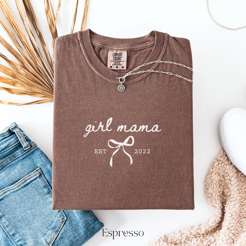May include: An espresso-colored short-sleeve t-shirt featuring the white text girl mama, a graphic of a bow, and the text EST 2022. The shirt has a Comfort Colors brand label at the inner neckline and a silver-toned chain necklace resting on it.